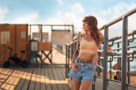 A young Caucasian tanned woman with tattoos poses on the embankment. A single stylish hipster girl...