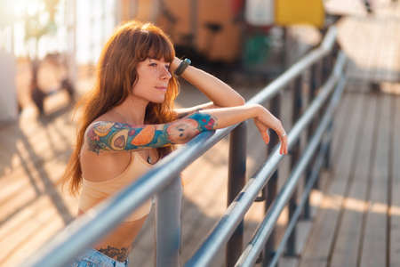 Portrait of young caucasian tanned woman with tattoos poses at fence. Alone stylish hipster girl...