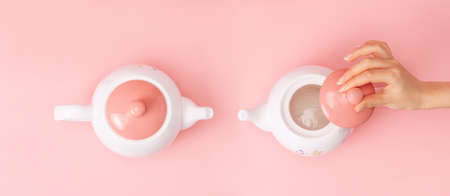 Banner. Two white porcelain teapots with a pink lid on a pink background. Female hand removes the lid from the kettle. flat lay. copyspace. Concept of Breast Cancer Awareness Month.の写真素材