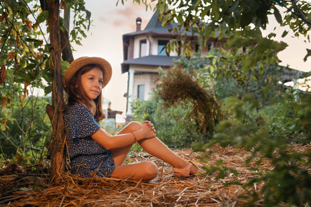 Summer organic gardening. Cute caucasian little girl wearing straw hat sits on the ground in garden. The concept of local farming and Earth Day.の写真素材