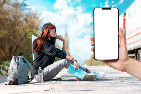 Hitchhiking. A caucasian woman in cap and sunglasses sitting along the road, holding a paper map and...