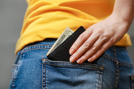 A woman's hand take out a black leather wallet with American dollars from the back pocket of her jeans. Close up, back view. The concept of salary, savings and wealth.の写真素材
