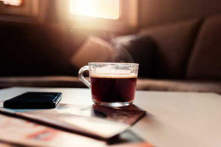 Glass cup of hot coffee on table in cozy living room. Window with morning sunlight and sofa in...