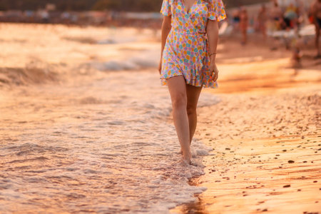 A slender woman in a summer dress walks along the sea surf near the shore. close-up. The golden sunset is reflected in the water. The concept of a summer vacation and smooth beautiful legs.の写真素材