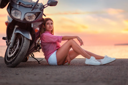 Caucasian young woman sitting and posing near motorcycle. Golden sunset and motorbike on the...