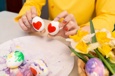Female hands holding two painted with heart shapes eggs near plate with easter decorations. Concept...