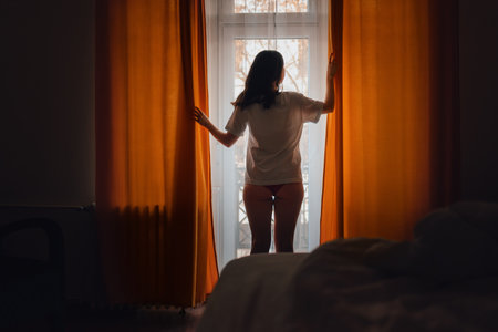 new day. Silhouette of a woman opening orange curtains in the morning at window. rear view. Concept of freedom, hope and happiness.の写真素材