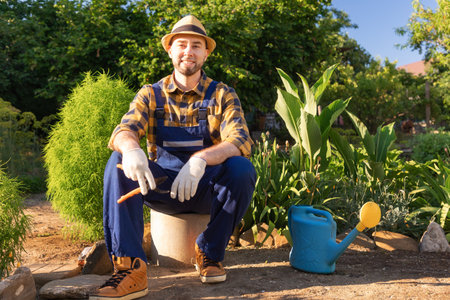 Happy bearded, handsome man in overalls and a straw hat sits and relax, holding a pruner. In the background is a backyard and a kitchen garden. The concept of home gardening.の写真素材