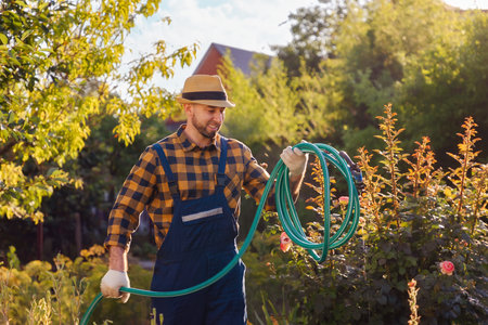 Happy bearded young handsome man in overalls and a straw hat, winding a hose for watering plants. In the background is a backyard and a garden. The concept of gardening and horticulture.の写真素材