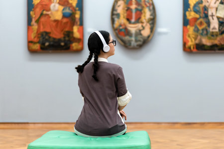 Back view of young Caucasian pretty woman wearing headphones and contemplates ancient artefacts....