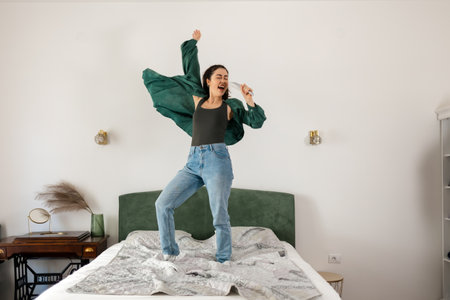 Winning casino bet. Young Caucasian happy woman is singing in hairbrush and dancing on bed. Concept of dance and vocals learning.の写真素材