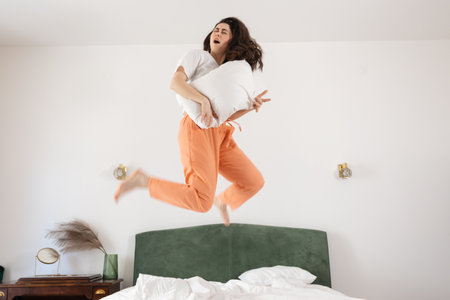 Happy young Caucasian woman funny jumping on bed and playing on air guitar. Bedroom interior. Concept of happiness and fun.の写真素材