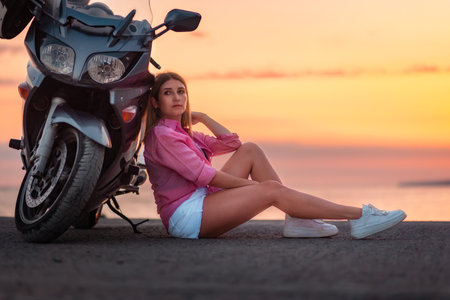 freedom. Pretty young woman sitting and posing near motorcycle. Golden sunset and motorbike on the background. copyspace. Concept of World Motorcyclist Day and moto trips.の写真素材