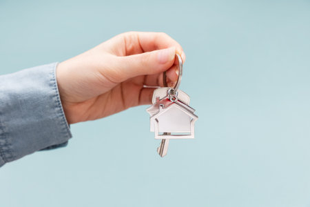 Female hand holding key and house keychain. Light blue background with copy space. Concept of mortgage, leasing of real estateの写真素材