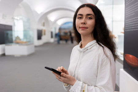 Portrait of young caucasian woman using smartphone in museum. light hall background. Concept of cultural exhibition and modern educational excursion.の写真素材