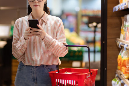 Close up of young Caucasian woman wearing glasses holds shopping basket and using smartphone. Concept of modern technology for shopping.の写真素材