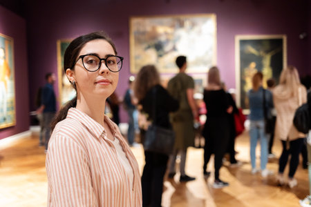 Portrait of young Caucasian woman wearing glasses. In background, people are looking at paintings. Concept of education and Museum Day.の写真素材