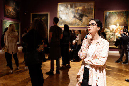 Thoughtful young Caucasian woman wearing glasses and looking at exhibition. In background, people are looking at paintings. Concept of Museum Dayの写真素材