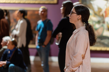 Side view of young Caucasian woman wearing glasses and looking at exhibition. People and masterpieces in the background. Concept of Museum Day.の写真素材