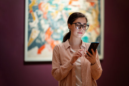 Young pretty Caucasian woman wearing glasses holds smartphone. Masterpieces in background. concept...