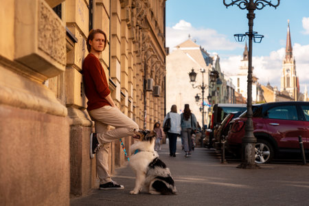 Friendship with pet. Side view of young handsome Caucasian man with pure breed aussie dog poses leans wall. Background of the city.の写真素材