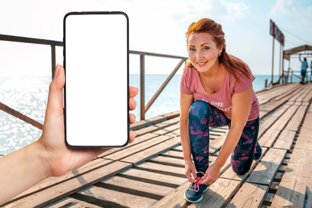 An adult smiling woman sitting down tying her shoelaces. In the background, the pier with the ship. Hand holding a smartphone mock up. The concept of sport and a healthy lifestyle.の写真素材