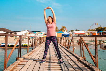 summer activities. An adult happy caucasian woman in sports clothes is doing sports on the pier by the sea. Sports and healthy lifestyle.の写真素材