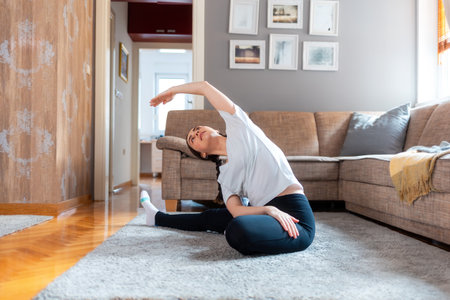 Young Caucasian woman sits on floor in living room and performs stretching bends. Home yoga and fitness.の写真素材