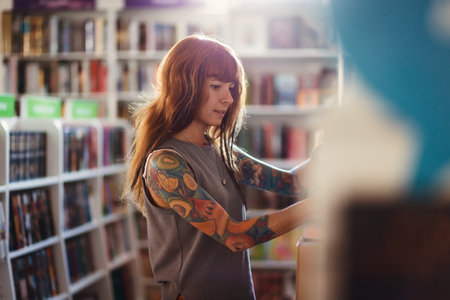 Young smiling Caucasian woman with tattoo choosing books in bookstore. view from shelf. Concept of education and shopping in book shop.の写真素材