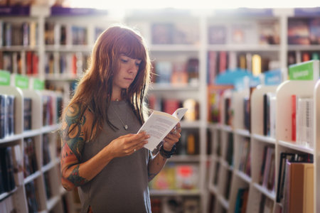 Side view of young Caucasian woman with tattoo posing reading book in bookstore. Concept of education and shopping.の写真素材