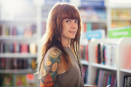 Portrait of young Caucasian smiling woman with tattoo posing in blurred background of bookstore. student buy textbook. concept of education.の写真素材