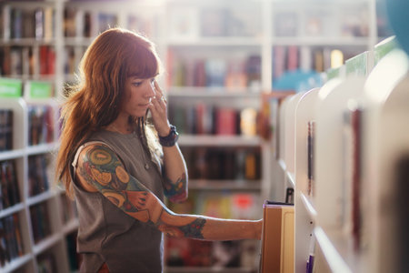 Young smiling Caucasian woman with tattoo choosing books in bookstore. View from shelf. Copy space. Concept of education and shopping in book shop.の写真素材
