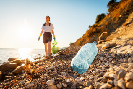 Defocused view of woman volunteer holds full plastic bag of trash and walking down of pebble wild beach. Close up of plastic bottle in foreground. Concept of protection ocean and Earth Day.の写真素材