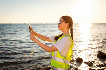 Inspection environmentalist. Side view of young Caucasian woman wearing glasses using tablet analyzing water in ocean. In background is ocean, coast and sunset. Concept of environmental protectionの写真素材