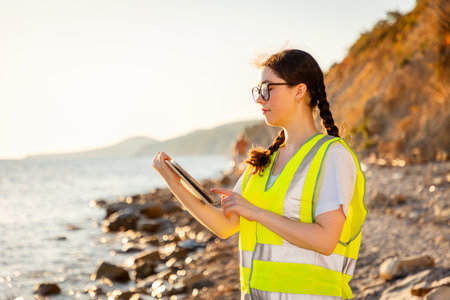 Inspection environmentalist. Side view of young Caucasian woman wearing glasses and vest using tablet for report. Concept of environmental protection of the ocean.の写真素材