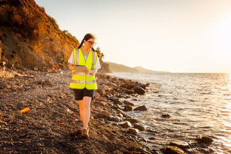 Volunteer woman wearing vest and using pad for analyzing sea water. In background is ocean, coast and sunset. Copy space. Concept of environmental pollution and ecology.の写真素材