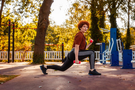 Side view of Caucasian pretty mid adult woman does squats and stretching using dumbbells and looking in camera. Fitness in city park.の写真素材