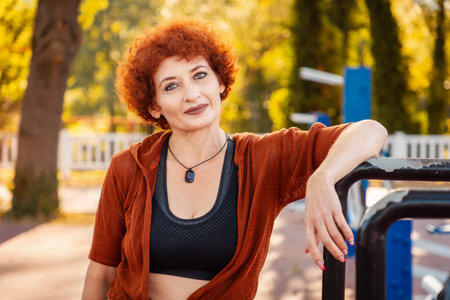 Portrait of adult Caucasian smiling woman with makeup and ginger curly hair looking in camera posing in sports park. Concept of healthy lifestyle and fitness.の写真素材