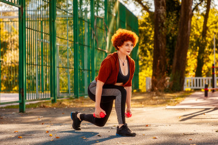 Adult Caucasian woman is doing sports in city park. Athlete does squats with dumbbells in her hands. Healthy lifestyle and fitness.の写真素材