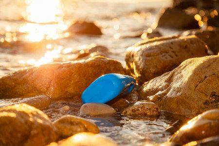 Close-up of blue plastic bottle lying in coastal rocks of sea. Sunset illuminates shore. Concept of environmental pollution.の写真素材