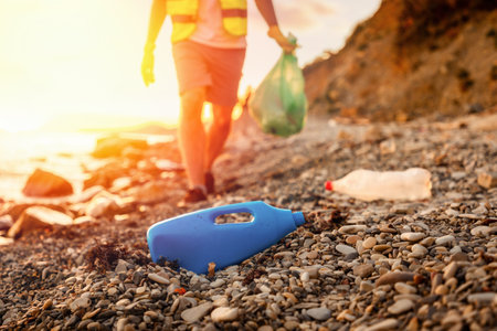 Defocused view of walking volunteer holding full plastic bag at pebble wild beach. Close up of plastic bottle in foreground. Concept of protection environment.の写真素材