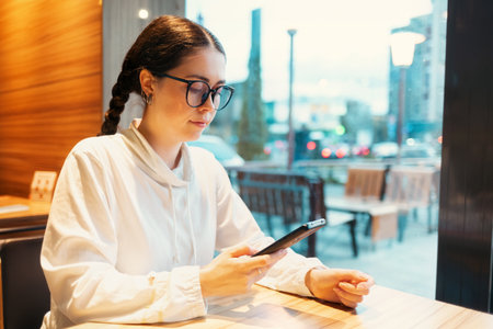 Freelance. Young Caucasian woman wearing glasses is sitting at table by window and using smartphone. Wi fi in cafes and coworking.の写真素材