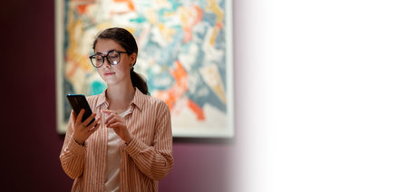 Web banner of Museum Day. Young pretty Caucasian woman wearing glasses holds smartphone. Masterpieces in the background. Mock up. Concept of cultural education.の写真素材