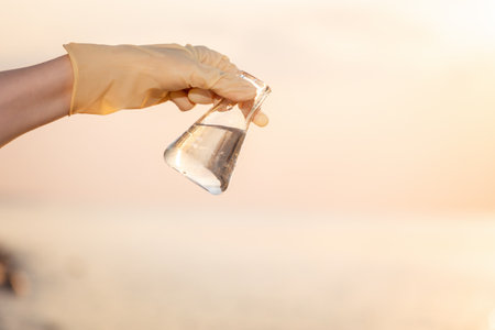 Close-up of hand wearing rubber glove shows glass flask with sample of pure water. Sunset sky and ocean on background. Copy space. Concept of science experiment and ecology.の写真素材