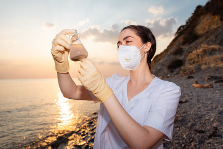 Portrait of woman scientist in rubber gloves and protective mask examines glass flask with sample of dirty water from sea. Ocean in background. Concept of environmental protection.の写真素材