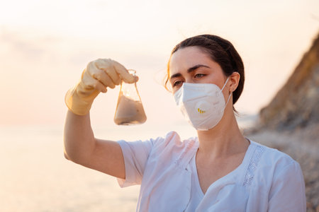 Portrait of woman ecologist wearing rubber gloves and protective mask examines glass flask with sample of dirty water from ocean. Concept of environmental protection and expertise.の写真素材