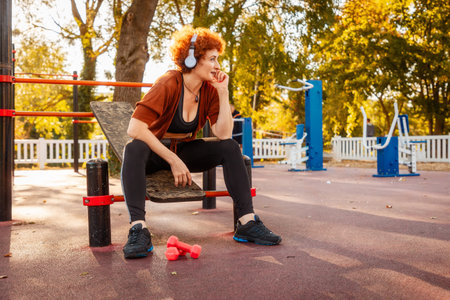 Mid adult Caucasian woman is sitting on playground and listening to music with headphones. Autumn...