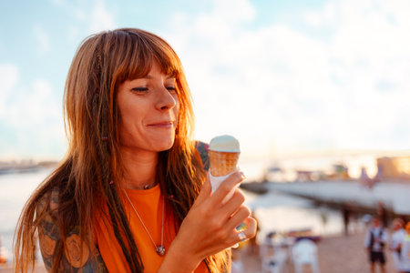 Portrait of young smiling happy Caucasian woman student with tattoos holds ice cream. Sunset beach in background. Concept of freedom and summer vacation.の写真素材