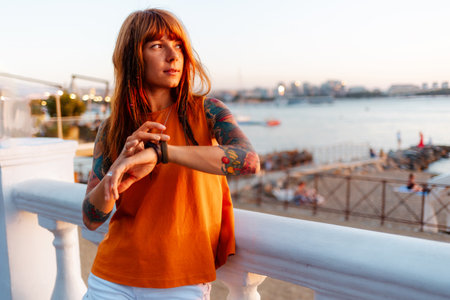 Young beautiful Caucasian woman with tattoos leaning on balustrade and looking on watch. Concept of waiting and psychology.の写真素材