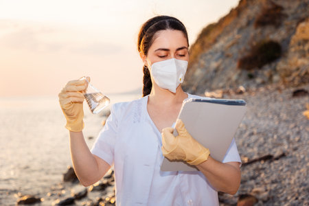 Portrait of woman scientist wearing rubber gloves and protective mask examines glass flask with sample of water from sea and holds tablet. Coast in background. Concept of recycle resources.の写真素材